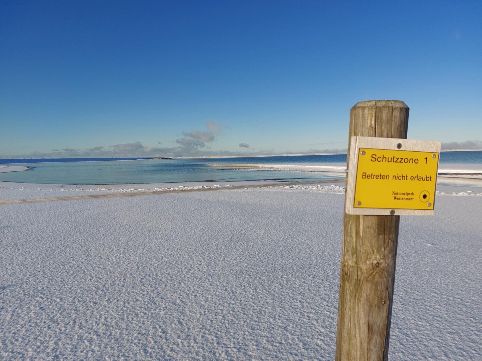 Holzpfahl mit gelbem Schild 'Schutzzone 1 Betreten nicht erlaubt Nationalpark Wattenmeer' vor verschneitem Strand und ruhiger See
