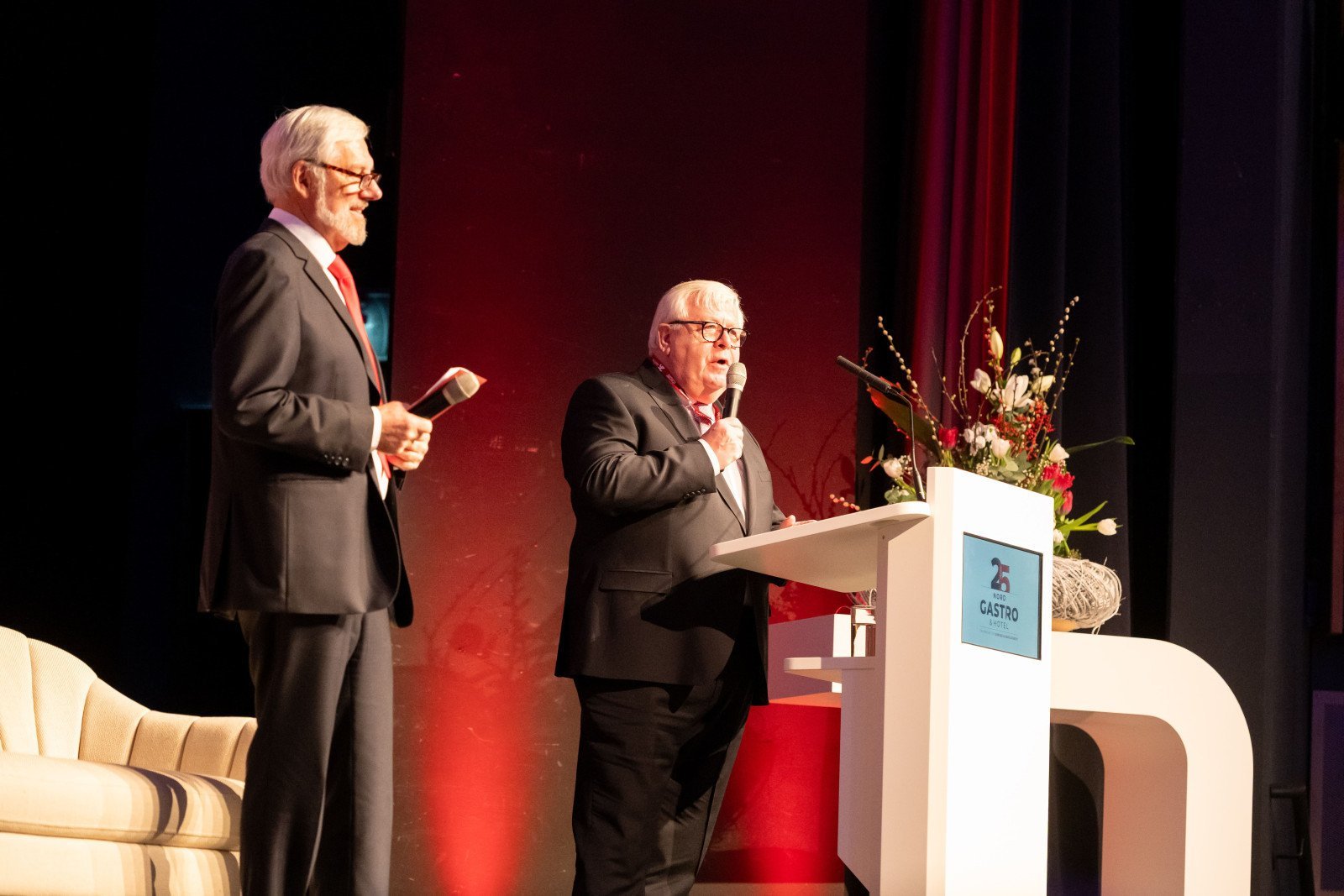 Zwei M&auml;nner in Anz&uuml;gen stehen auf einer B&uuml;hne, einer spricht am Rednerpult mit Blumenarrangement und Logo mit Schriftzug 'GASTRO'