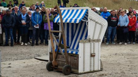 Ein weiß-blau gestreifter Strandkorb auf einem Holzwagen vor einer Menschenmenge im Freien.