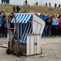 Ein weiß-blau gestreifter Strandkorb auf einem Holzwagen vor einer Menschenmenge im Freien.