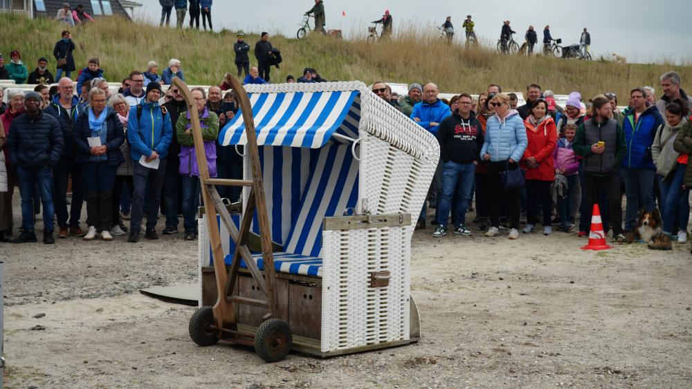 Ein weiß-blau gestreifter Strandkorb auf einem Holzwagen vor einer Menschenmenge im Freien.