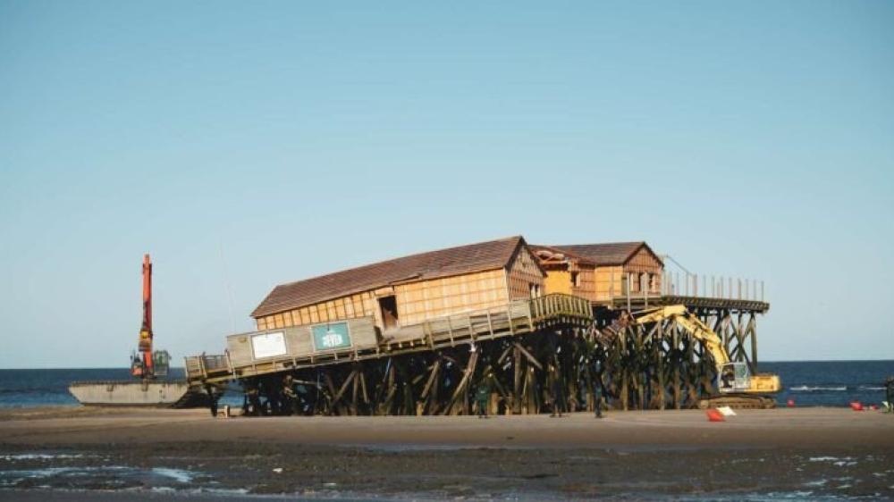 Ein Teil der Stelzen eines Pfahlbaus in St. Peter-Ording ist eingebrochen. Links und rechts neben dem Pfahlbau sind Bagger. Im Hintegrund die Nordsee und ein blauer Himmel.