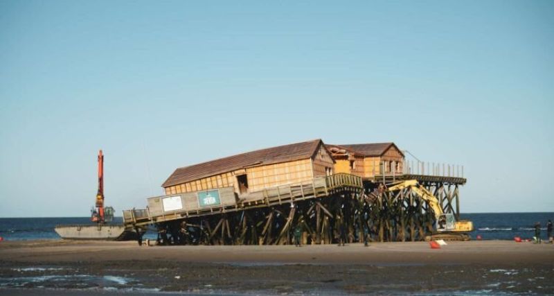 Ein Teil der Stelzen eines Pfahlbaus in St. Peter-Ording ist eingebrochen. Links und rechts neben dem Pfahlbau sind Bagger. Im Hintegrund die Nordsee und ein blauer Himmel.