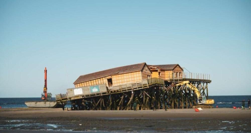 Ein Teil der Stelzen eines Pfahlbaus in St. Peter-Ording ist eingebrochen. Links und rechts neben dem Pfahlbau sind Bagger. Im Hintegrund die Nordsee und ein blauer Himmel.
