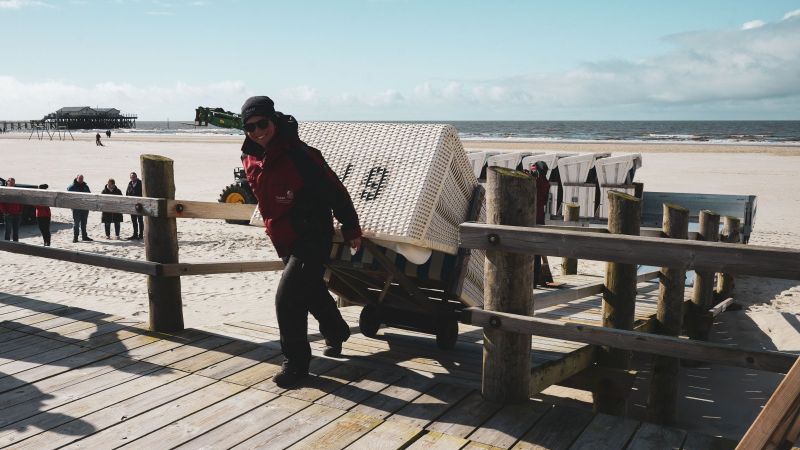 Eine Frau in roter Jacke und schwarzer Hose zieht einen Strandkorb auf ein Holzpodest