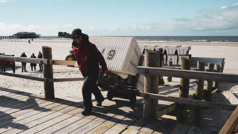 Eine Frau in roter Jacke und schwarzer Hose zieht einen Strandkorb auf ein Holzpodest
