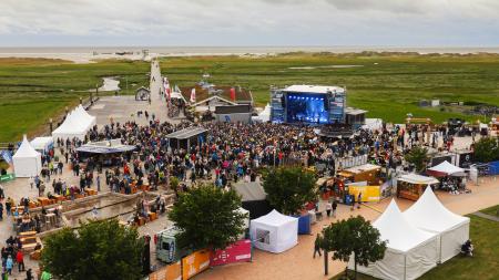 St. Peter-Ording Nachhaltigkeits-Festival. Zu sehen Eine Bühne auf einem Platz, im Hintergrund Seewiesen und weißer Sandstrand mit Pfahlbauten.
