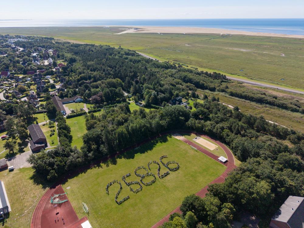 Menschen bilden auf dem Sportplatz der Nordseeschule St. Peter-Ording die Postleitzahl. Das Bild zeigt sie aus der Vogelperspektive. Im Hintergrund Wald, Häuser, Deich, Salzwiesen, weißer Strand und Meer.