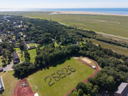 Menschen bilden auf dem Sportplatz der Nordseeschule St. Peter-Ording die Postleitzahl. Das Bild zeigt sie aus der Vogelperspektive. Im Hintergrund Wald, Häuser, Deich, Salzwiesen, weißer Strand und Meer.