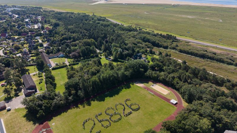 Menschen bilden auf dem Sportplatz der Nordseeschule St. Peter-Ording die Postleitzahl. Das Bild zeigt sie aus der Vogelperspektive. Im Hintergrund Wald, Häuser, Deich, Salzwiesen, weißer Strand und Meer.