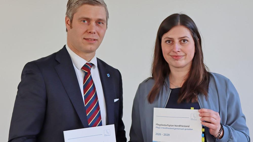 Zwei Personen stehen nebeneinander, die rechte hält ein Dokument mit Text, die linke trägt Anzug mit gestreifter Krawatte und Anstecknadel.