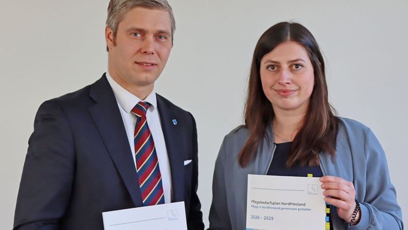 Zwei Personen stehen nebeneinander, die rechte hält ein Dokument mit Text, die linke trägt Anzug mit gestreifter Krawatte und Anstecknadel.