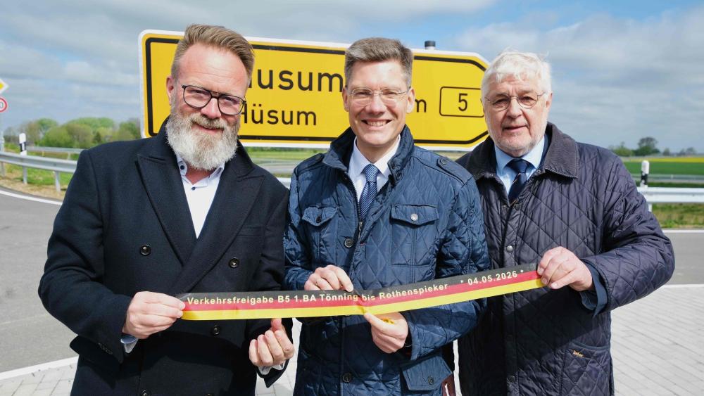 Drei Männer stehen vor einem gelben Verkehrsschild, welches mit Husum und einer 5 beschriftet ist
