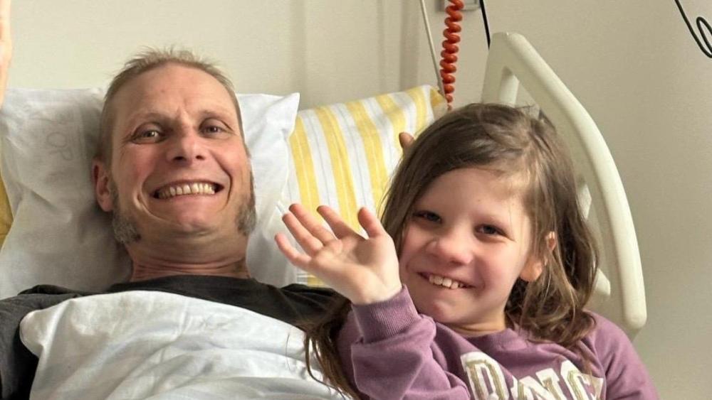 Ein Mann liegt in einem Krankenhausbett, neben ihm sitzt ein kleines Mädchen. Beide lächeln und winken in die Kamera.