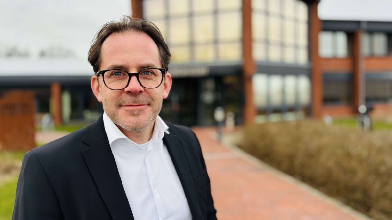 Mann in dunklem Anzug und weißem Hemd steht vor einem modernen Bürogebäude mit großen Fenstern und rotem Backstein