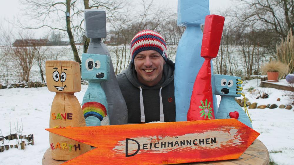 Ein Mann in einem Garten mit bunten Holzmännchen im Vordergrund. Im Garten liegt Schnee.