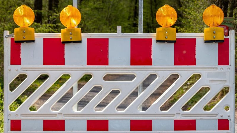 Absperrung mit rot-weißen Streifen und vier gelben Warnleuchten vor bewaldetem Hintergrund.