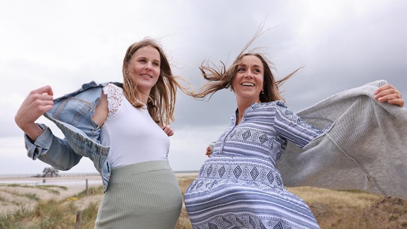 Zwei Frauen mit wehenden Jacken stehen an einem Strand mit Dünen und bewölktem Himmel