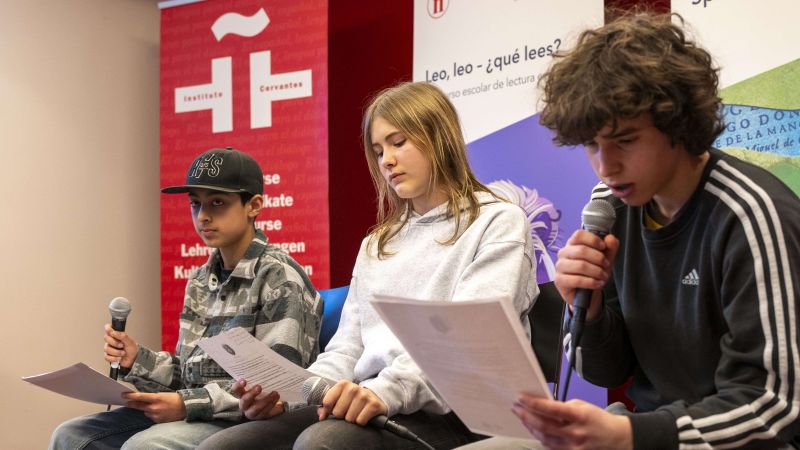 Drei Jugendliche sitzen nebeneinander auf Stühlen, zwei halten Mikrofone und alle drei halten Blätter in den Händen, im Hintergrund Banner mit spanischem Text