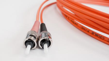 Nahaufnahme von zwei nicht eingesteckten metallischen Steckern eines orangen Glasfaserkabels, das auf einem weißen Untergrund liegt. Im Hintergrund ist das restliche Kabel unscharf aufgerollt.