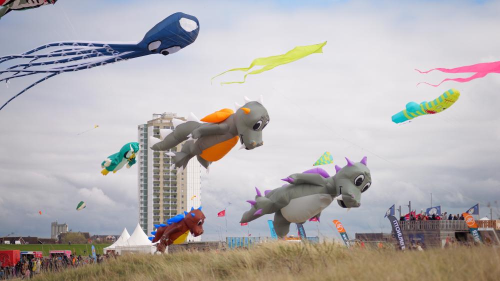Bunte Drachen am Himmel: Im Hintegrund ein Hochhaus, dass vor einem grünen Seedeich steht.