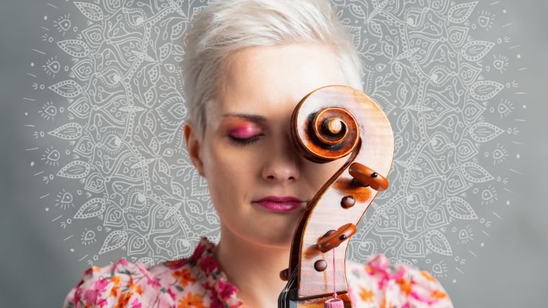 Frau mit kurzen blonden Haaren und pinkem Lidschatten hält ein Cello vor ihr Gesicht vor einen hellen Hintergrund mit Mandala-Muster.