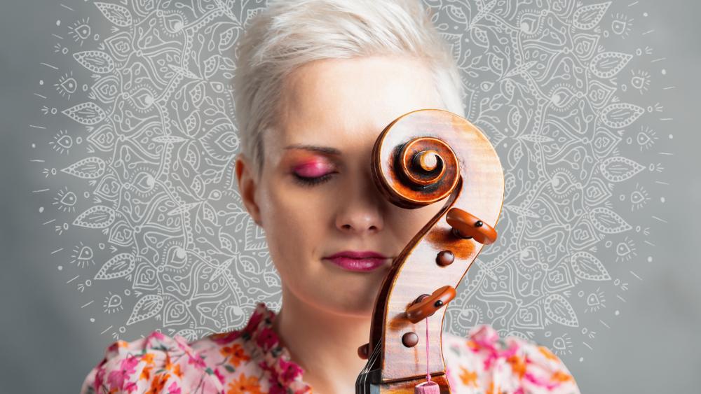 Frau mit kurzen blonden Haaren und pinkem Lidschatten hält ein Cello vor ihr Gesicht vor einen hellen Hintergrund mit Mandala-Muster.