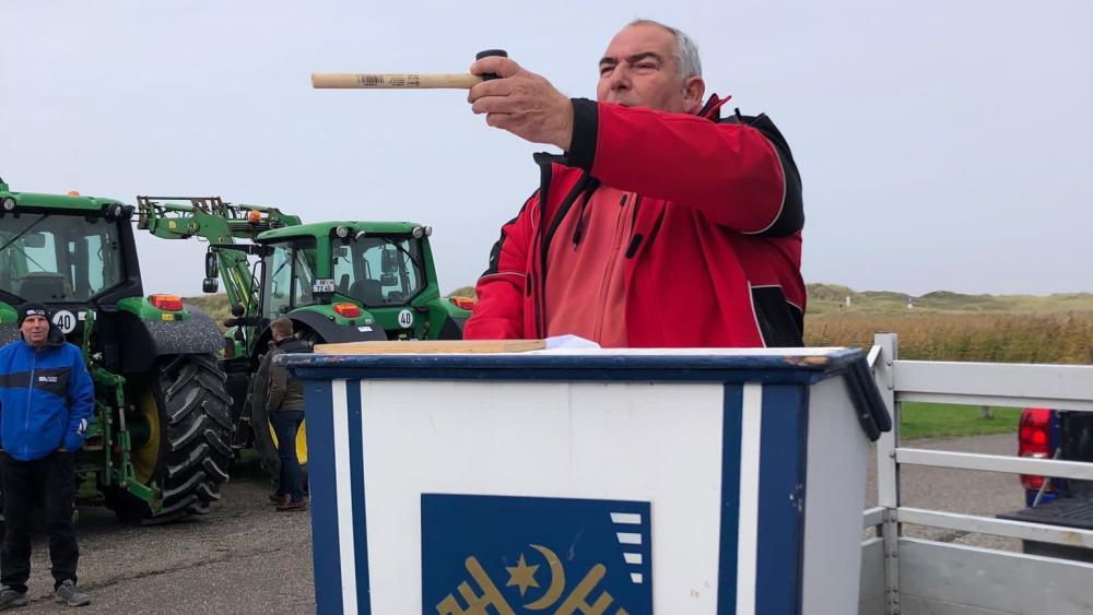 Ein Mann steht an einem Pult mit dem Gemeindesiegel St. Peter-Ordings. Im Hintergrund Dünen und zwei grüne Traktoren.