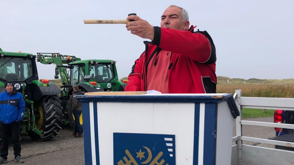 Ein Mann steht an einem Pult mit dem Gemeindesiegel St. Peter-Ordings. Im Hintergrund Dünen und zwei grüne Traktoren.