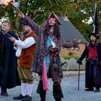 Vier Männer in historischen Kostümen, einer in Piratenkleidung mit gezückter Pistole und gezücktem Säbel, stehen auf einem gepflasterten Platz vor einem Haus mit Gartenzaun.