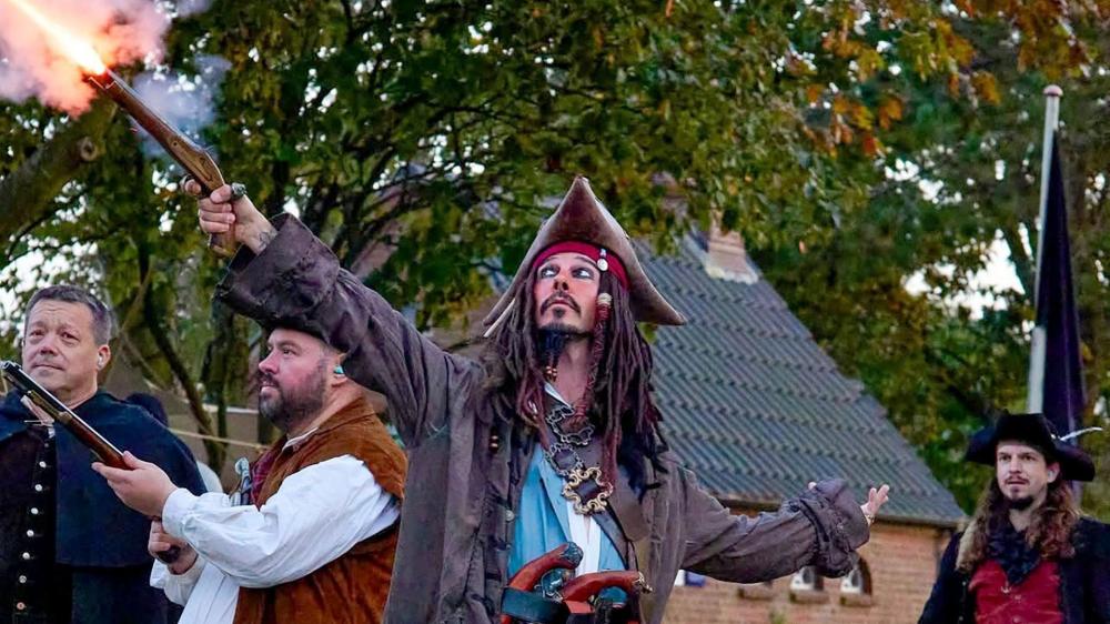 Vier Männer in historischen Kostümen, einer in Piratenkleidung mit gezückter Pistole und gezücktem Säbel, stehen auf einem gepflasterten Platz vor einem Haus mit Gartenzaun.