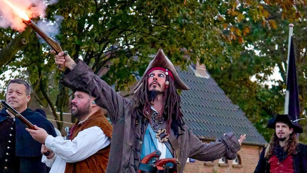 Vier Männer in historischen Kostümen, einer in Piratenkleidung mit gezückter Pistole und gezücktem Säbel, stehen auf einem gepflasterten Platz vor einem Haus mit Gartenzaun.