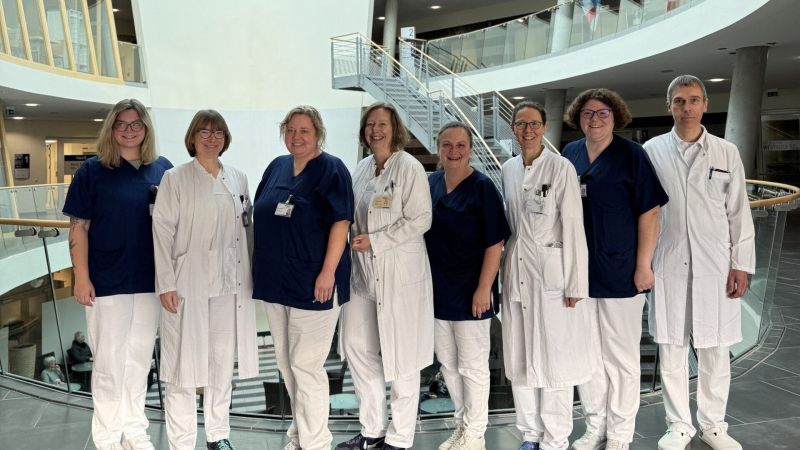 Gruppe von acht Personen in medizinischer Kleidung steht in einem modernen Gebäude mit Treppe und Glasgeländer im Hintergrund