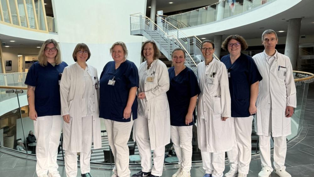 Gruppe von acht Personen in medizinischer Kleidung steht in einem modernen Gebäude mit Treppe und Glasgeländer im Hintergrund