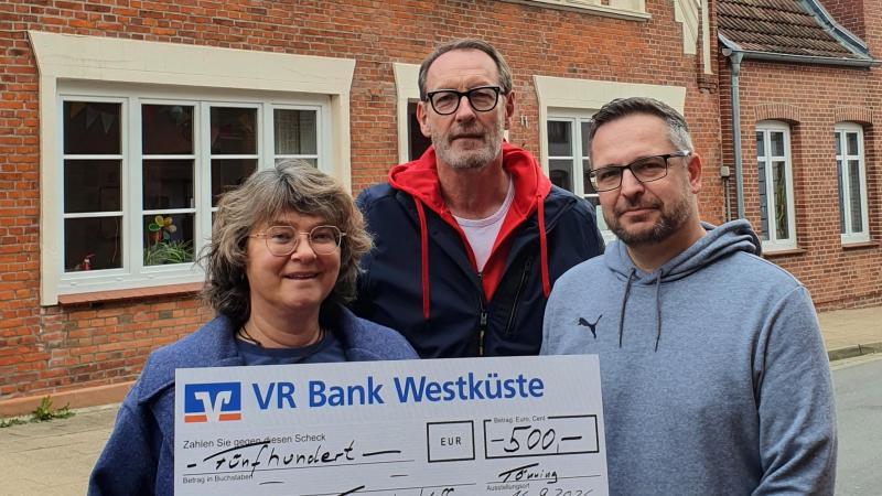 Drei Personen vor einem Backsteinhaus halten einen großen Scheck mit der Aufschrift 'VR Bank Westküste' und dem Betrag '500 EUR'.