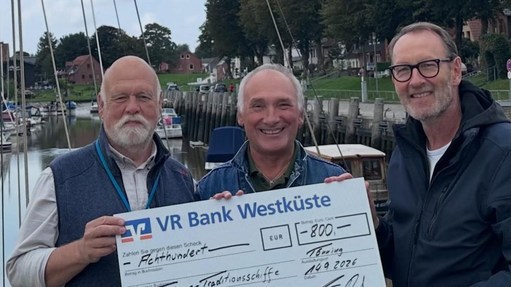 Drei Männer stehen am Hafen, zwei halten einen großen Scheck der VR Bank Westküste über 800 Euro für Tönninger Traditionsschiffe