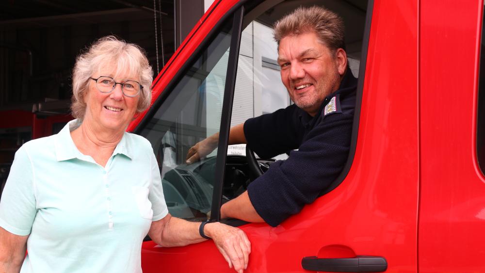 Eine Frau lehnt an ein rotes Feuerwehrauto der Feuerwehr Oldenswort. Im Auto sitzt ein Mann.