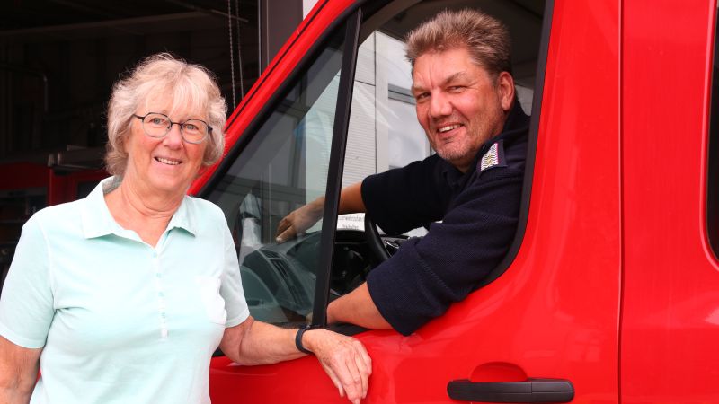 Eine Frau lehnt an ein rotes Feuerwehrauto der Feuerwehr Oldenswort. Im Auto sitzt ein Mann.