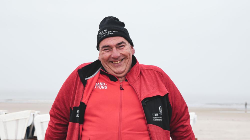 Ein Mann in roter Jacke lehnt lächelnd an einem Holzgeländer. Im Hintergrund weißer Sandstrand.