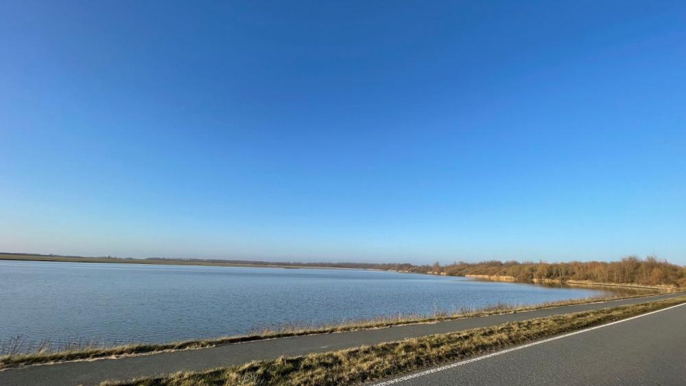 Blick auf einen ruhigen See mit Uferweg und angrenzender Straße unter klarem Himmel.