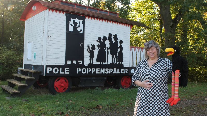 Eine Frau in gepunktetem Kleid steht mit Handpuppe vor einem Bauwagen. Auf dem Bauwagen steht Pole Poppenspäler