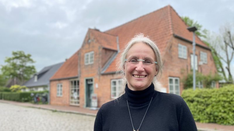Eine Frau mit Brille steht lächelnd vor einem Backsteinhaus.