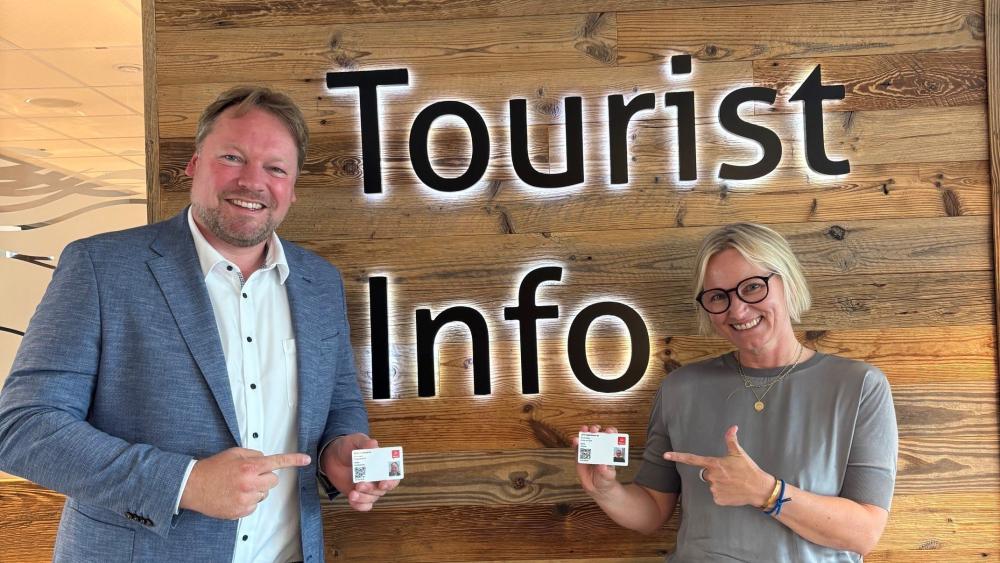Ein Mann und eine Frau stehen vor einer Holzwand, auf der Touristinfo steht.