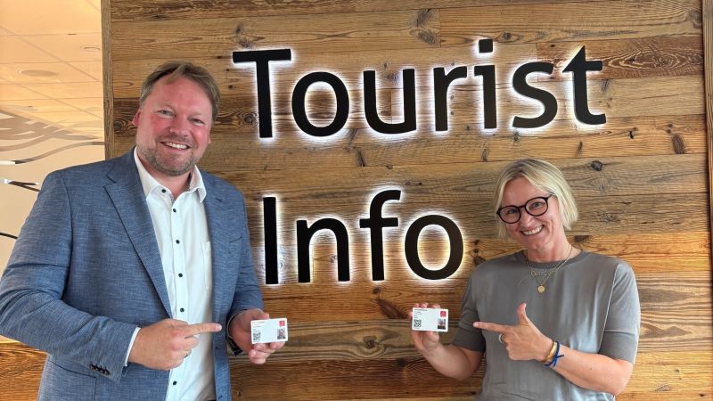 Ein Mann und eine Frau stehen vor einer Holzwand, auf der Touristinfo steht.