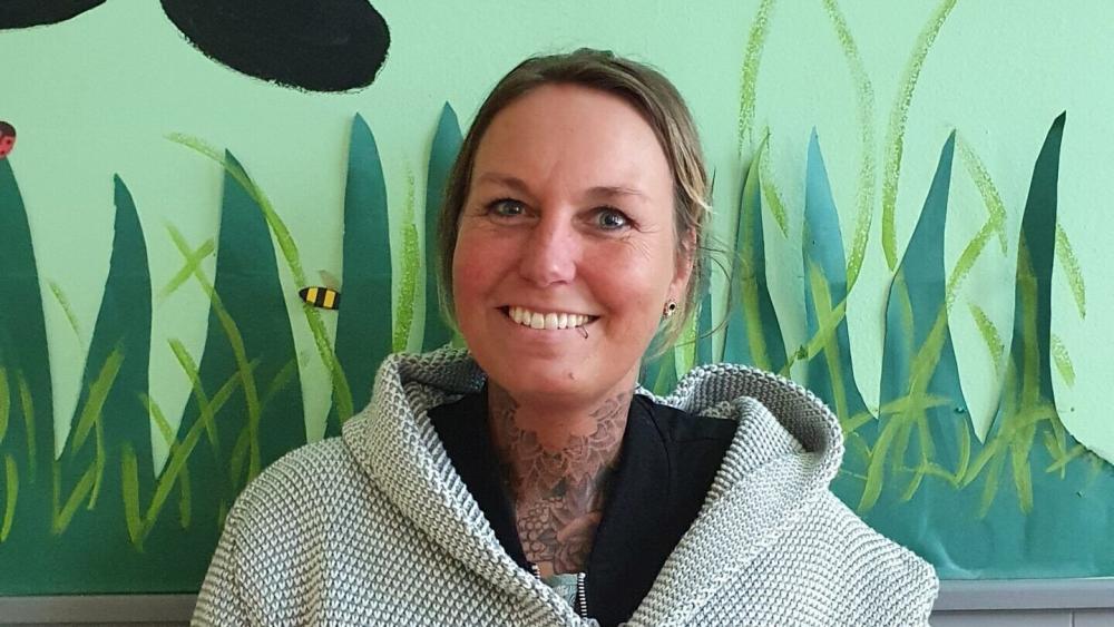 Person mit Kapuzenjacke und sichtbarem Hals-Tattoo vor einer Wand mit großflächigem Pflanzen- und Insektenmotiv