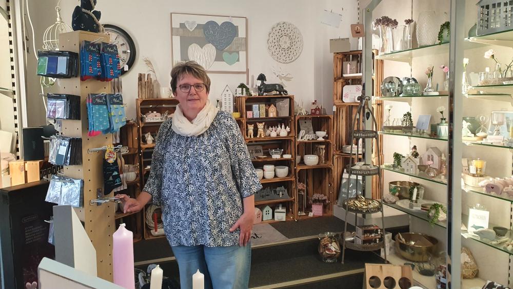 Frau mit Brille und weißem Schal steht in einem Laden mit Regalen voller Dekorationsartikel.
