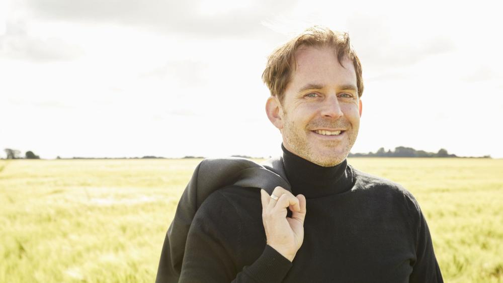 Ein in schwarz gekleideter Mann, dessen Oberkörper zu sehen ist, steht lächelnd in einem Feld.
