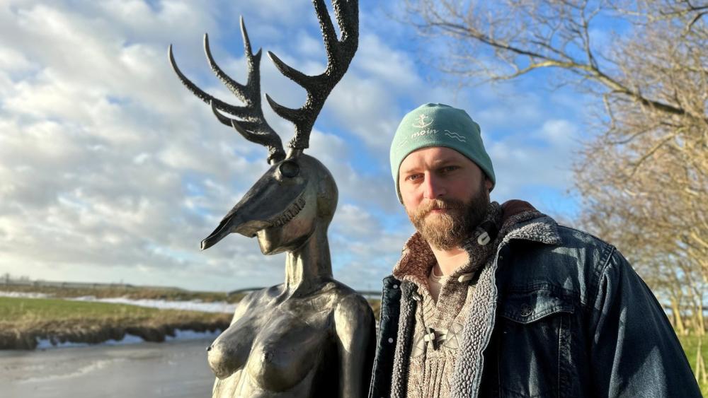 Bronzefigur mit Hirschgeweih und weiblichem Oberkörper neben einer Person in Jeansjacke vor Fluss und blauem Himmel