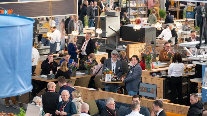 Menschen stehen und sitzen an Tischen und Theken in einer belebten Messehalle mit moderner Einrichtung und Dekoration.
