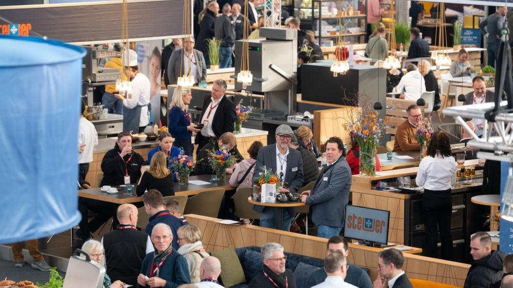 Menschen stehen und sitzen an Tischen und Theken in einer belebten Messehalle mit moderner Einrichtung und Dekoration.